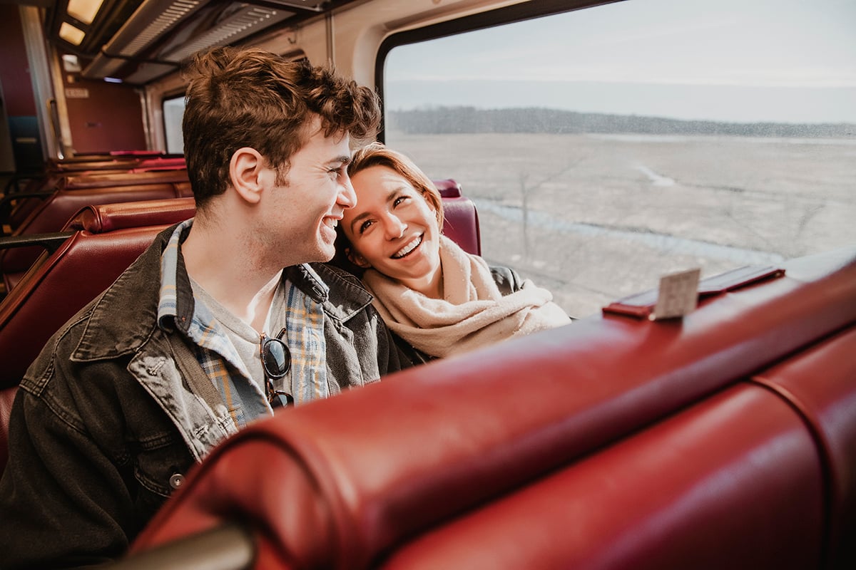 A smiling man and women ride the train next to a window featuring seascapes.